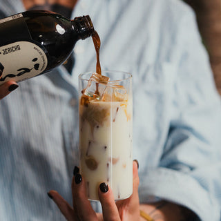 A person in a light blue button-up shirt pours a dark brown liquid from a bottle into a tall glass of iced milk, with the bottle label partially visible.