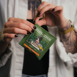 A close-up of a person holding a green packet of 'Jeeves + Jericho' Mojito Mint tea. The person has tattoos on their arms, wears a white button-down shirt, and a gold watch.