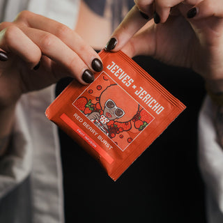 A close-up of a person holding a single Jeeves + Jericho Red Berry Blast tea packet. The person has dark brown manicured nails and is wearing a white button-down shirt over a black top.