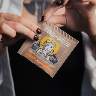 A close-up of a person holding a single Jeeves & Jericho Chamomile & Lavender tea packet. The person has dark brown nail polish and is wearing a white shirt over a black top.