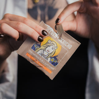 A close-up of a person's hands with dark brown nail polish tearing open a single-serve tea packet from the brand Teepot & Jericho. The packet is labeled Chamomile & Lavender and features an illustration of a woman with a sun behind her head.
