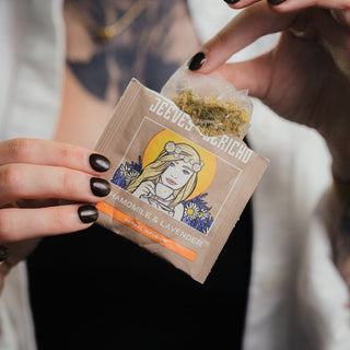 A close-up of a person holding a tea bag package labeled 'Jeeves Herbd' and 'Chamomile & Lavender,' with a tea bag being pulled open to show the herbal tea inside. The person has dark nail polish and a white shirt, with a black top underneath.