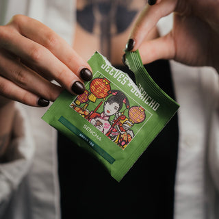 A close-up of a person holding a green Jeeves + Jericho Sencha single-serve packet, showing the illustrated packaging and the person's hands with dark nail polish.